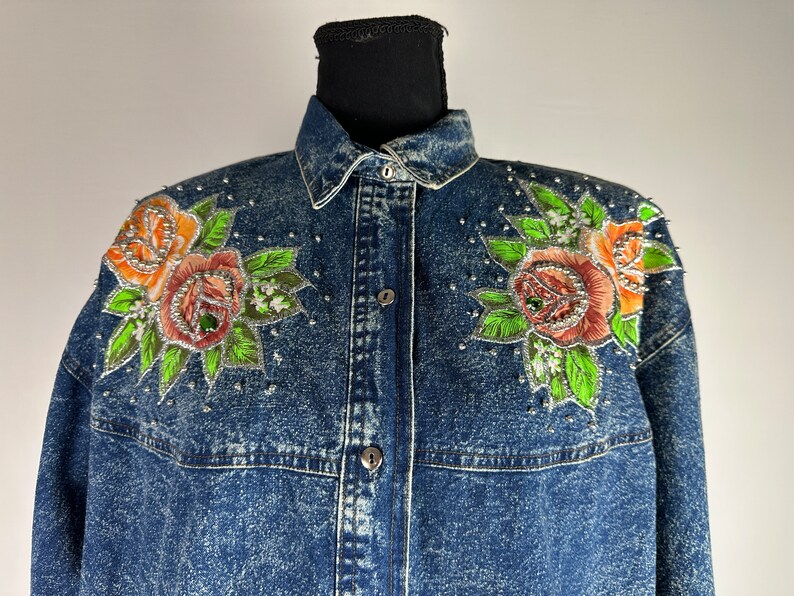 以下が含まれることがあります： オレンジ、ピンク、グリーンのバラが刺繍された、ブルーのデニムジャケット。シルバーの装飾が施されています。