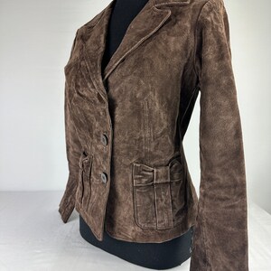May include: A brown suede jacket with a bow detail on the pocket. The jacket has a double breasted closure and long sleeves.