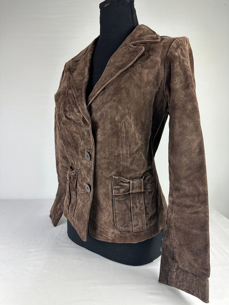 May include: A brown suede jacket with a bow detail on the pocket. The jacket has a double breasted closure and long sleeves.