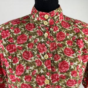Pode incluir: Uma camisa branca de botões com colarinho alto e mangas curtas. A camisa é coberta por uma estampa de rosas vermelhas em um fundo verde.