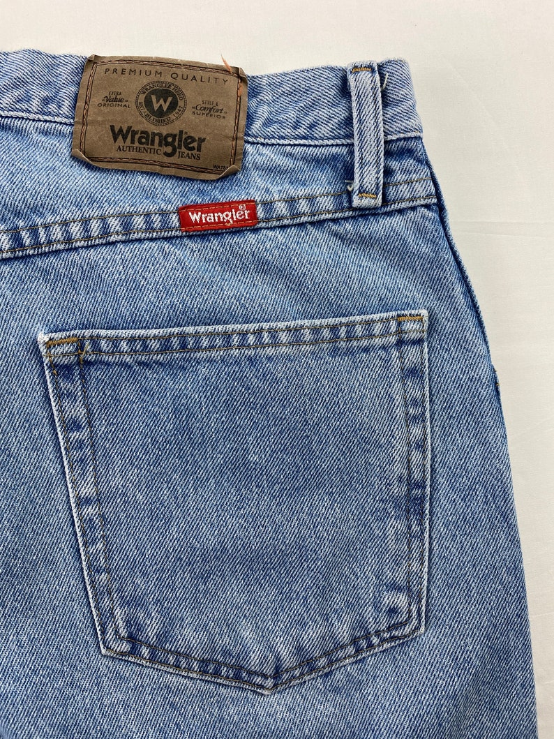 May include: Light blue denim jeans with a back pocket and a brown leather Wrangler label that says "Premium Quality" and "Authentic Jeans".
