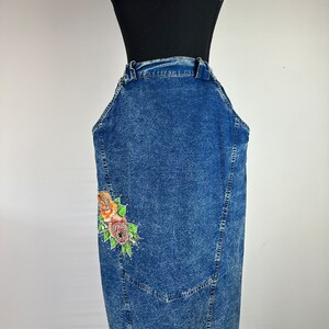 以下が含まれることがあります： サイドに花柄の刺繍が施された青いデニムのオーバーオールスカート。スカートはハイウエストでストレートフィットです。
