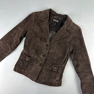 May include: A brown suede jacket with a button closure and two pockets with bows on the front. The jacket has a label that says "XOXO" on it.