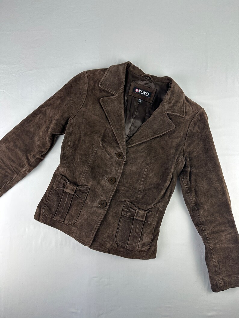 May include: A brown suede jacket with a button closure and two pockets with bows on the front. The jacket has a label that says "XOXO" on it.