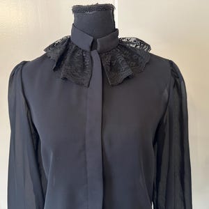 K&ouml;nnte beinhalten: Eine schwarze Bluse mit langen &Auml;rmeln und einem Spitzenkragen. Der Kragen ist breit, gekr&auml;uselt und hat einen gewellten Rand.