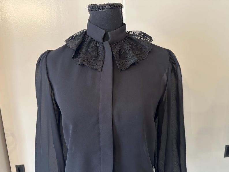 K&ouml;nnte beinhalten: Eine schwarze Bluse mit langen &Auml;rmeln und einem Spitzenkragen. Der Kragen ist breit, gekr&auml;uselt und hat einen gewellten Rand.