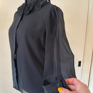 K&ouml;nnte beinhalten: Eine schwarze, durchsichtige Bluse mit Spitzenkragen und langen &Auml;rmeln. Die &Auml;rmel haben eine geraffte Detail am &Auml;rmelabschluss.