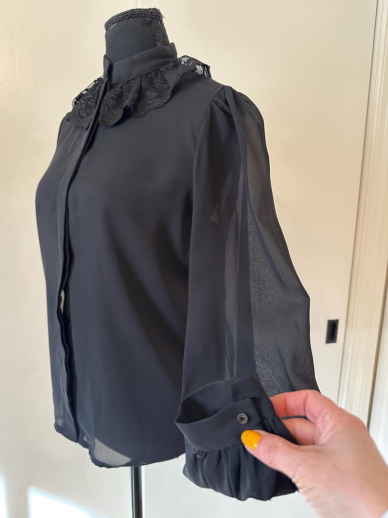 K&ouml;nnte beinhalten: Eine schwarze, durchsichtige Bluse mit Spitzenkragen und langen &Auml;rmeln. Die &Auml;rmel haben eine geraffte Detail am &Auml;rmelabschluss.
