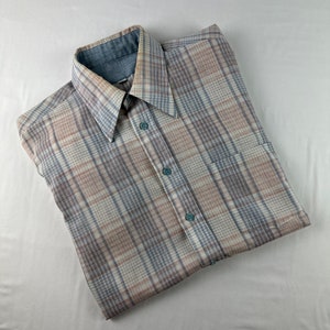 May include: A folded button-down shirt with a plaid pattern in shades of beige, blue, and brown. The shirt has a wide collar and a single pocket. The buttons are a light blue color.