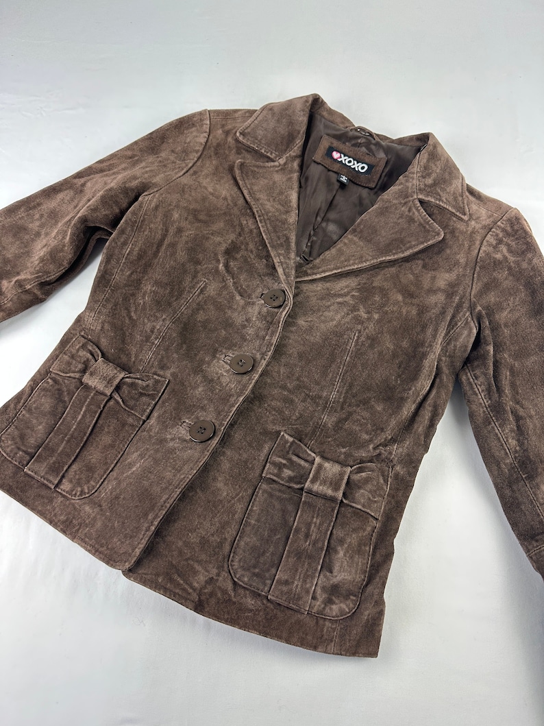 May include: A brown suede jacket with a button closure and two pockets with bow details. The jacket has a label that says "XOXO".
