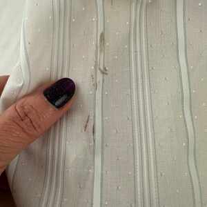 May include: Close-up of a white fabric with thin gray stripes and a small white dot pattern. There are a few brown stains on the fabric.