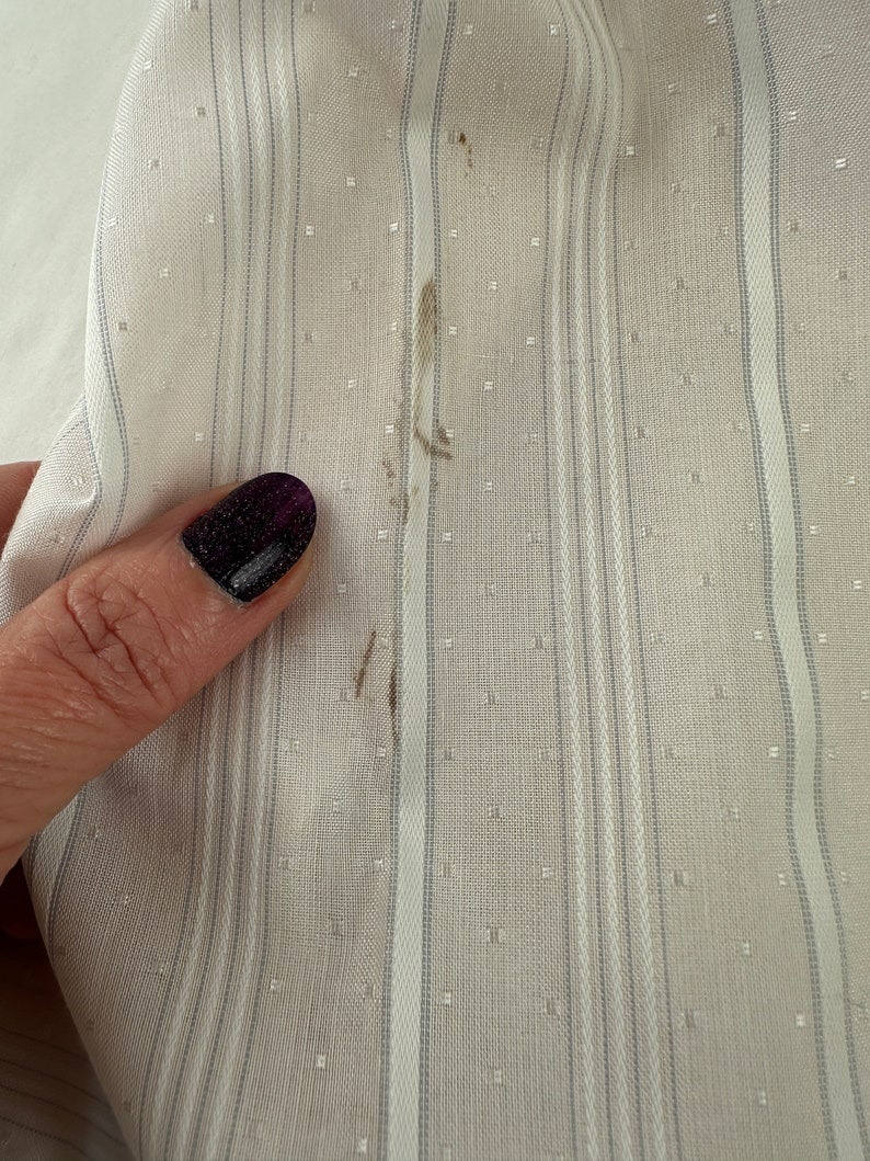 May include: Close-up of a white fabric with thin gray stripes and a small white dot pattern. There are a few brown stains on the fabric.