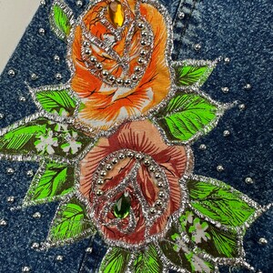 以下が含まれることがあります： 2つの刺繍されたバラ、オレンジとピンクのバラが、銀色の縁取りとラインストーンで飾られたデニムジャケット。バラは緑の葉と白い花に囲まれています。