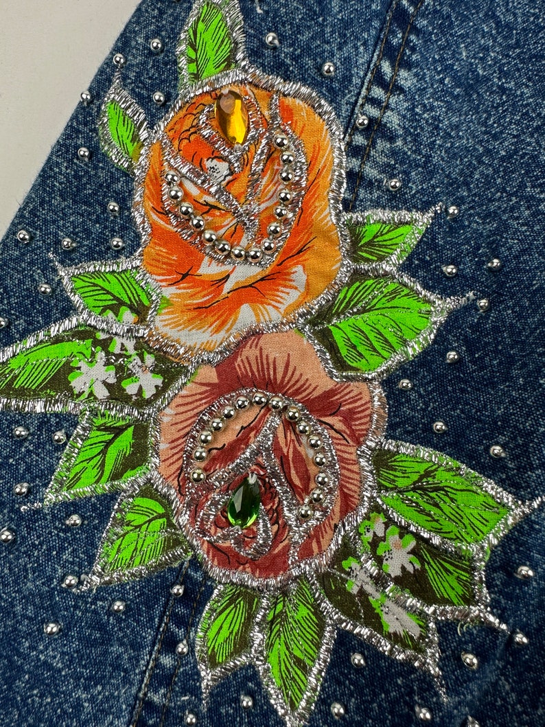 以下が含まれることがあります： 2つの刺繍されたバラ、オレンジとピンクのバラが、銀色の縁取りとラインストーンで飾られたデニムジャケット。バラは緑の葉と白い花に囲まれています。