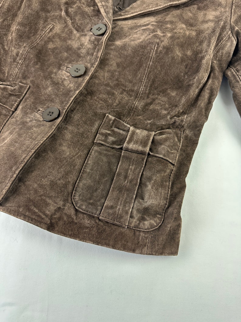 May include: A brown suede jacket with a bow detail on the front pocket and three buttons down the front.