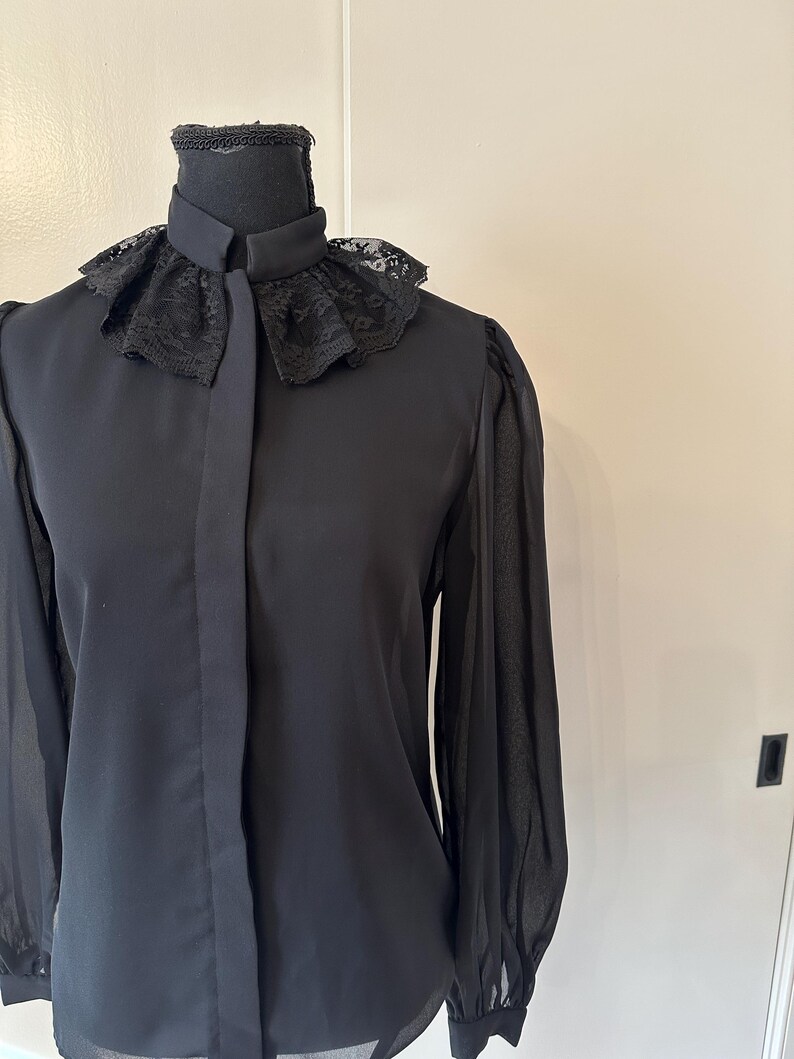 K&ouml;nnte beinhalten: Eine schwarze Bluse mit langen &Auml;rmeln und einem Spitzenkragen. Die Bluse hat einen Knopfleisten vorne und durchsichtige &Auml;rmel.