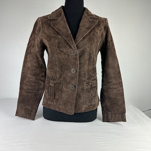 May include: A brown suede jacket with a button closure and pockets. The jacket has a classic style and is perfect for layering.