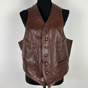 May include: A brown leather vest with a V-neck and multiple button closures. The vest has two flap pockets on the front.