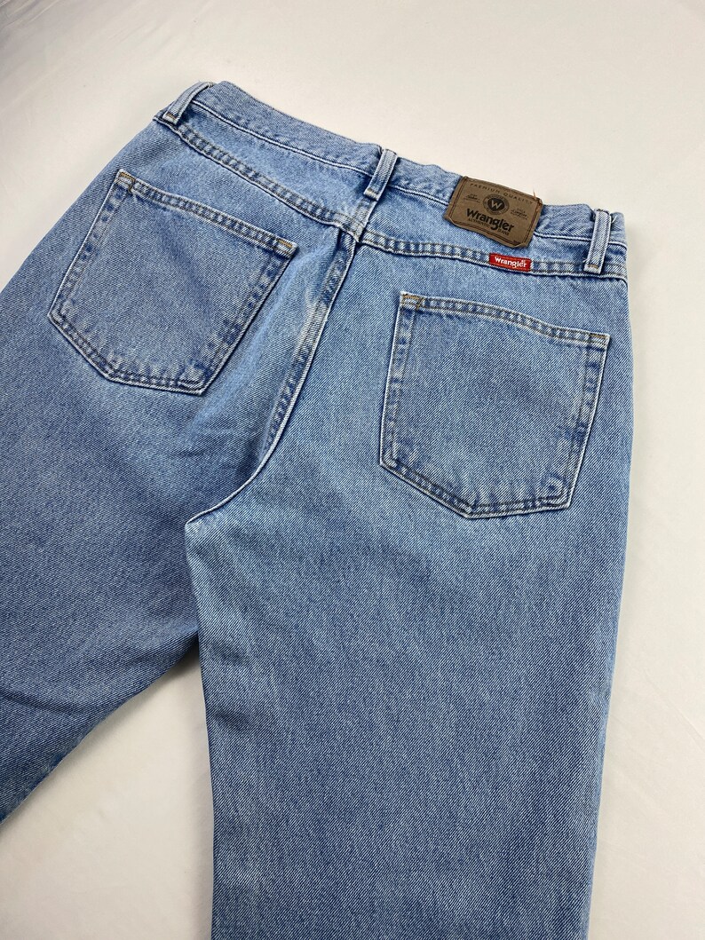 May include: A pair of light blue denim jeans with a brown leather Wrangler label on the back pocket. The label has the Wrangler logo and the text "Premium Quality".