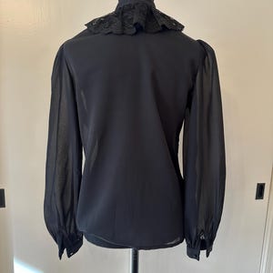 K&ouml;nnte beinhalten: Eine schwarze, durchsichtige Bluse mit einem Spitzenkragen. Die Bluse hat lange &Auml;rmel und eine figurbetonte Silhouette.