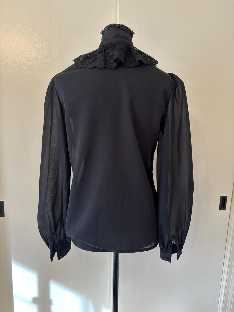 K&ouml;nnte beinhalten: Eine schwarze, durchsichtige Bluse mit einem Spitzenkragen. Die Bluse hat lange &Auml;rmel und eine figurbetonte Silhouette.