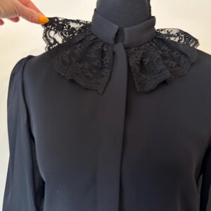 K&ouml;nnte beinhalten: Eine schwarze Bluse mit langen &Auml;rmeln und einem Spitzenkragen. Der Kragen ist gekr&auml;uselt und hat ein zartes Muster.