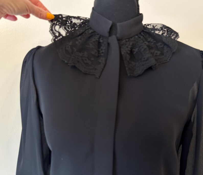 K&ouml;nnte beinhalten: Eine schwarze Bluse mit langen &Auml;rmeln und einem Spitzenkragen. Der Kragen ist gekr&auml;uselt und hat ein zartes Muster.