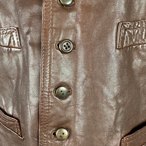 May include: Close-up of a brown leather vest with four brown buttons down the front.