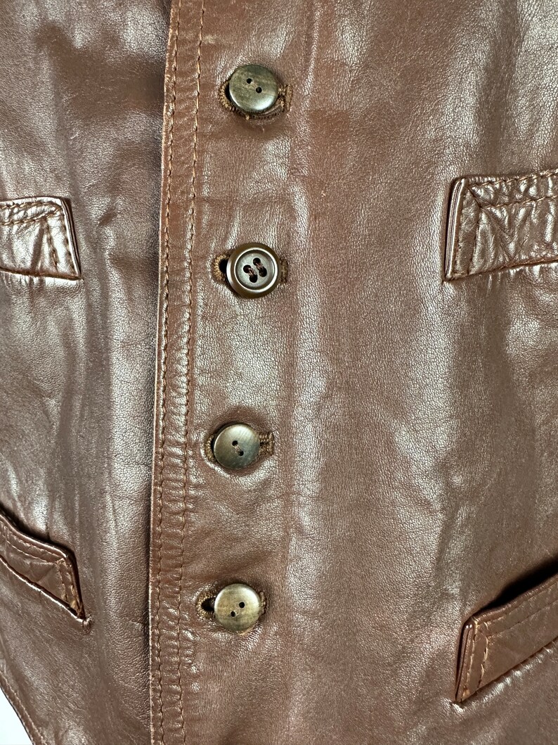 May include: Close-up of a brown leather vest with four brown buttons down the front.