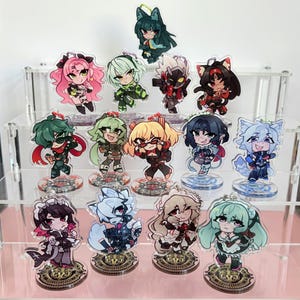May include: A collection of 14 acrylic character stands featuring anime-style characters. The characters are in various poses and have different colored hair, clothing, and accessories. The stands are on a clear acrylic base with a gold design.
