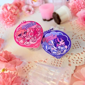 Pode incluir: Dois recipientes circulares de plástico com tampas destacáveis com personagens estilo anime e o texto Heaven's Sweetest. Uma tampa é rosa com uma personagem em traje branco, a outra é roxa com uma personagem em traje azul e branco.
