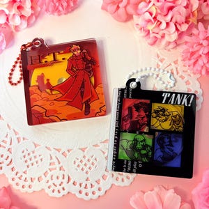 May include: Two square acrylic keychains. The left one features a character in a red coat with the text H.T. Humanoid Typhoon. The right one is black with the text TANK! and four square panels showing illustrated characters in red, yellow, green, and blue.