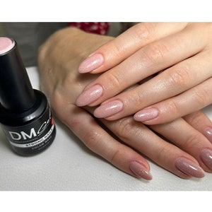 Puede incluir: Primer plano de una mano con esmalte de uñas de purpurina rosa. La mano sostiene una botella de esmalte de uñas etiquetada como "DM Strong Nail Color Base".