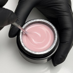 May include: A jar of light pink nail gel is being stirred with a brush. The gel is in a black container with a silver rim. The person stirring the gel is wearing black gloves.