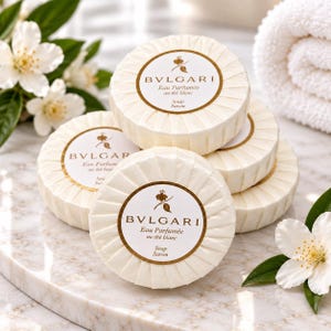 Puede incluir: Una pila de cinco jabones redondos envueltos en papel blanco plisado con la etiqueta Bvlgari Eau Parfumée au thé blanc. Cada jabón presenta una etiqueta circular con borde dorado, un pequeño icono de flor, la marca y el texto Soap Savon.