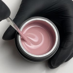 Puede incluir: Un frasco de gel de uñas rosa claro se agita con un pequeño aplicador. El gel está en un recipiente negro con un borde plateado. La persona que agita el gel lleva un guante negro.