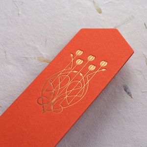 Può includere: Un segnalibro rosso con un design floreale dorato. Il design presenta un fiore stilizzato con uno stelo e foglie.