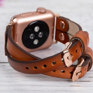 May include: A brown leather Apple Watch band with a rose gold buckle. The band is double-wrapped and has a classic look.