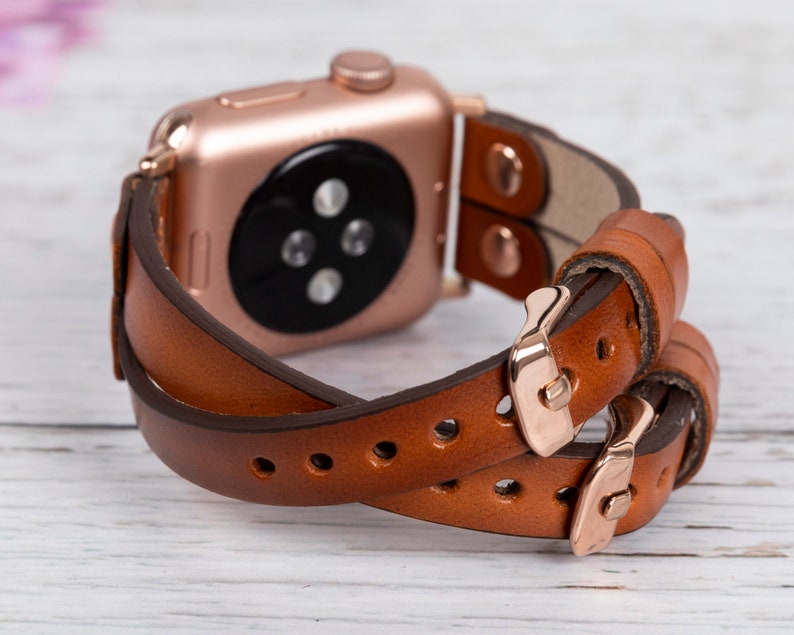 May include: A brown leather Apple Watch band with a rose gold buckle. The band is double-wrapped and has a classic look.