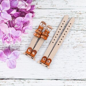 May include: Two brown leather watch bands with gold clasps and a white background. The bands have small holes along the edges.