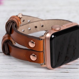 May include: A rose gold Apple Watch with a brown leather band. The band is double layered and has a knotted design.