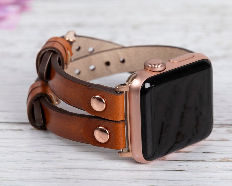 May include: A rose gold Apple Watch with a brown leather band. The band is double layered and has a knotted design.
