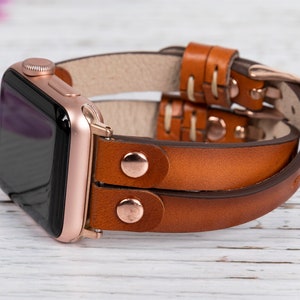 May include: A brown leather Apple Watch band with a rose gold buckle. The band is double-layered and has a stitched design.