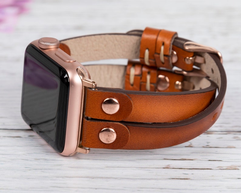 May include: A brown leather Apple Watch band with a rose gold buckle. The band is double-layered and has a stitched design.