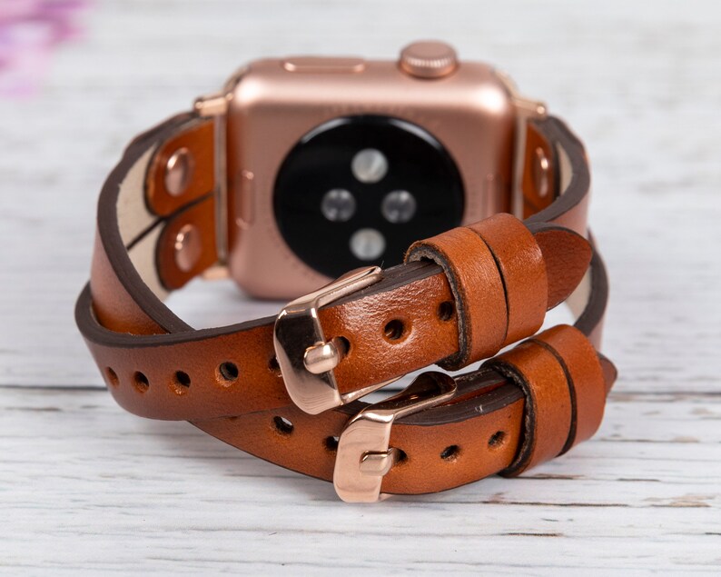 May include: A brown leather double wrap watch band with rose gold hardware. The band is designed to fit Apple Watches.