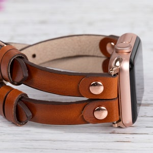 May include: A brown leather double wrap watch band with rose gold hardware and a rose gold Apple Watch.