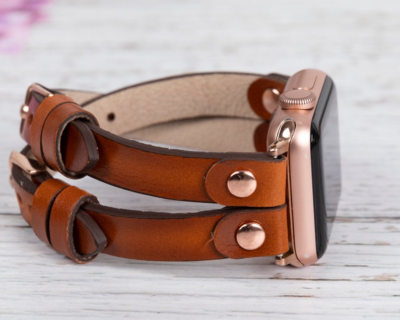 May include: A brown leather double wrap watch band with rose gold hardware and a rose gold Apple Watch.