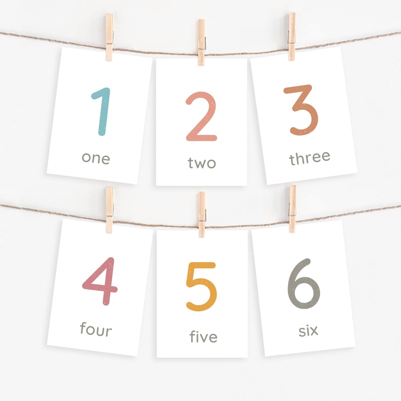 Peut inclure: Six cartes blanches suspendues &agrave; une ficelle par des pinces &agrave; linge en bois. Chaque carte affiche un chiffre de 1 &agrave; 6 dans des couleurs vari&eacute;es, ainsi que son &eacute;quivalent en lettres. Les chiffres sont en bleu, p&ecirc;che, marron, rose, jaune et gris.