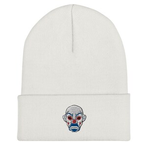 K&ouml;nnte beinhalten: Wei&szlig;er Beanie mit einem gestickten Clown-Gesicht. Das Clown-Gesicht hat rote Augen, einen blauen Mund und rote Wangen. Die M&uuml;tze ist aus weichem Strickmaterial.