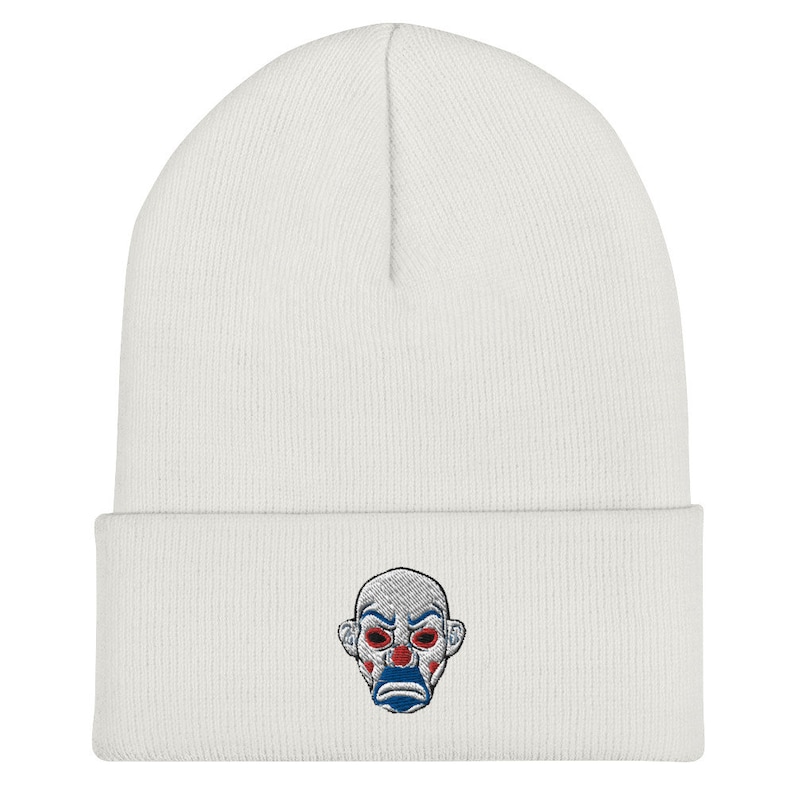 K&ouml;nnte beinhalten: Wei&szlig;er Beanie mit einem gestickten Clown-Gesicht. Das Clown-Gesicht hat rote Augen, einen blauen Mund und rote Wangen. Die M&uuml;tze ist aus weichem Strickmaterial.